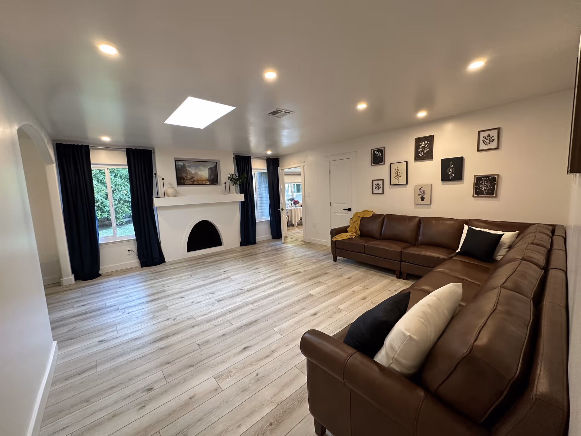 A spacious living room with light wood flooring, white walls, and a white ceiling with recessed lighting and a skylight. There is a large brown leather sectional sofa with black and white pillows on the right side. The far wall features a white fireplace with a curved opening, flanked by two windows with dark curtains. Above the fireplace is a framed landscape painting and decorative items. Several framed botanical prints are arranged on the wall above the sofa. A doorway leads to another room in the background.