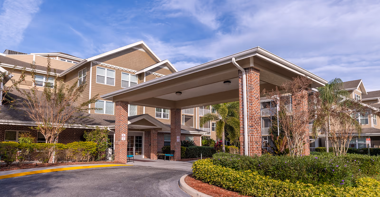 Exterior view of a senior living facility with a covered entrance supported by brick columns, surrounded by landscaped greenery and trees under a partly cloudy sky.