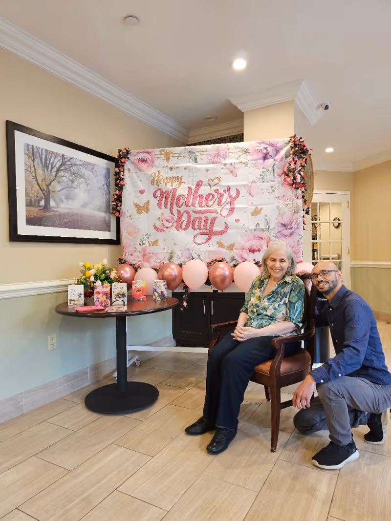 An elderly woman sitting on a chair next to a man kneeling beside her in a decorated room. Behind them is a floral banner with the words 'Happy Mother's Day' surrounded by pink and rose gold balloons. A round table nearby holds several greeting cards and a bouquet of flowers. The room has beige walls, a framed picture of a tree-lined path, and tiled flooring.