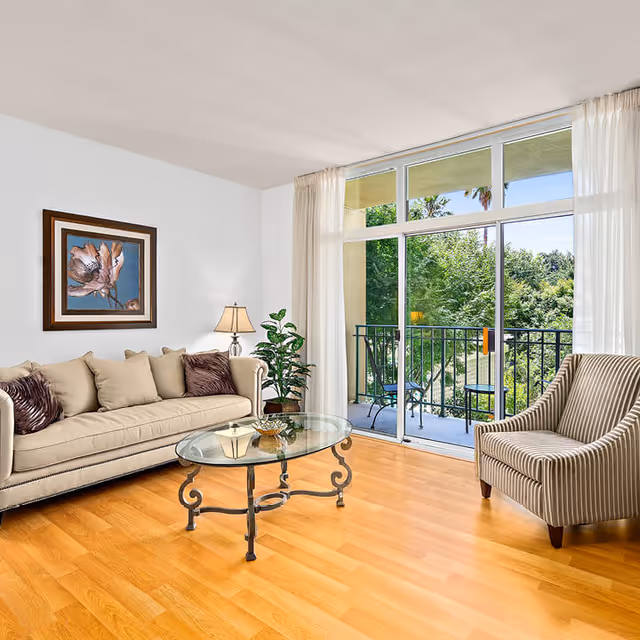 Bright living room with a beige sofa, glass coffee table, striped armchair, potted plant, and sliding glass doors leading to a balcony.