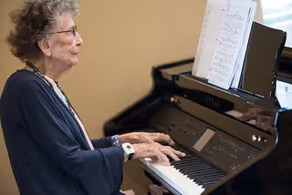 An elderly woman with glasses playing a black digital piano with sheet music placed on the music stand in a well-lit room.