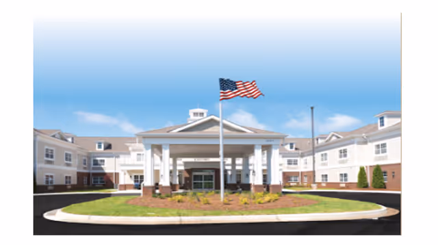 Front exterior view of The Goldton at Athens senior living facility with a circular driveway, a covered entrance supported by white columns, and an American flag flying on a flagpole in front of the building under a clear blue sky.