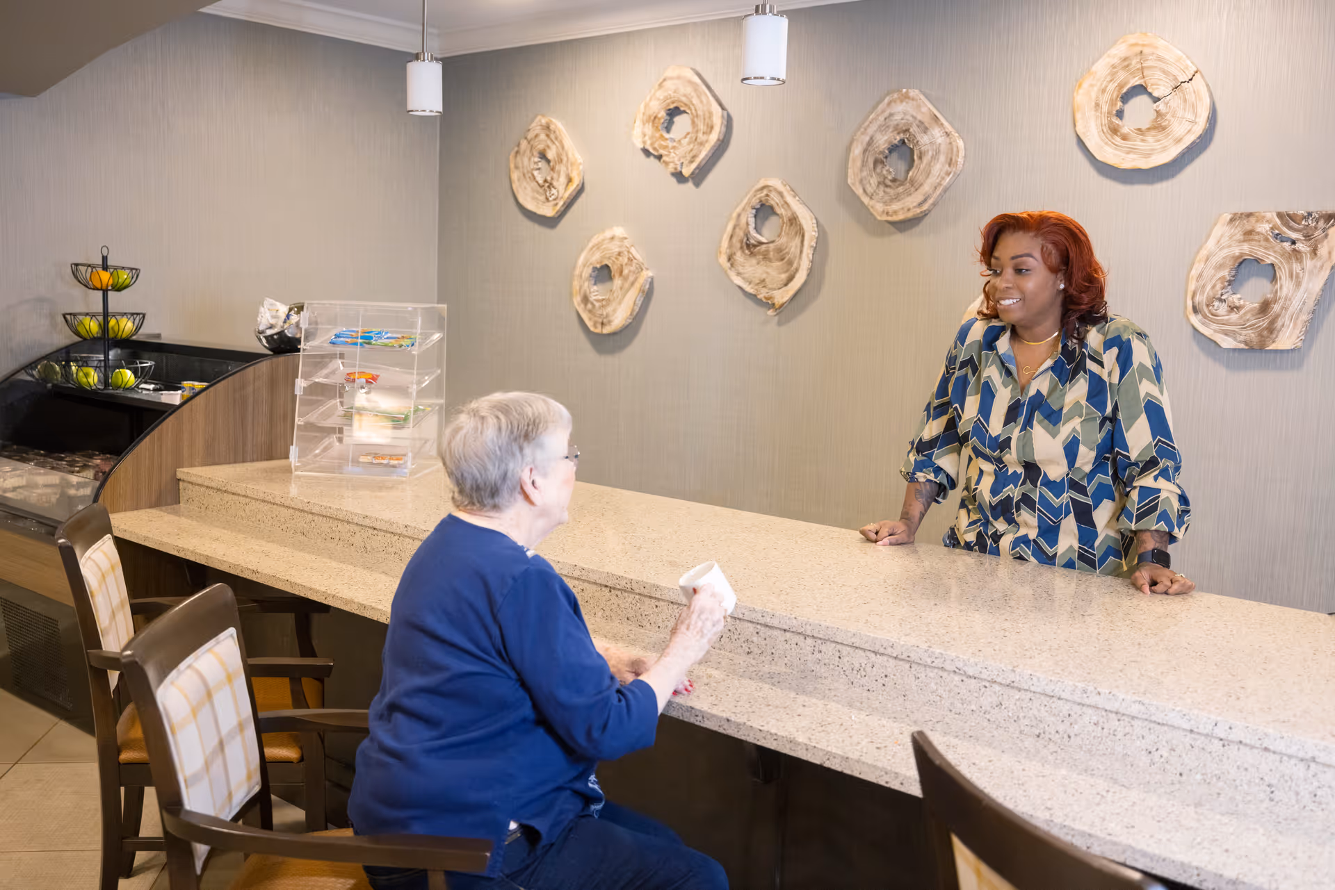 An elderly woman sitting at a counter holding a cup, talking to a smiling staff member standing behind the counter in a senior living facility. The background features decorative wooden wall art and a display case with fruit and snacks.