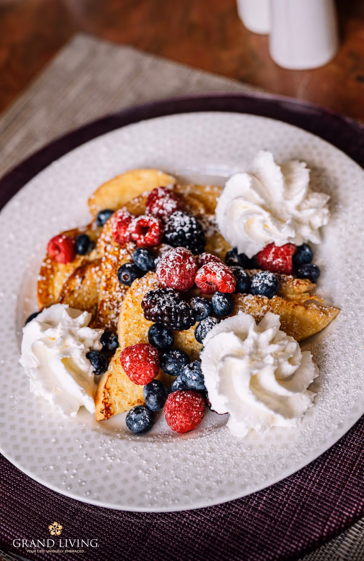 A plate of French toast topped with fresh raspberries, blackberries, blueberries, powdered sugar, and three dollops of whipped cream, placed on a purple placemat.