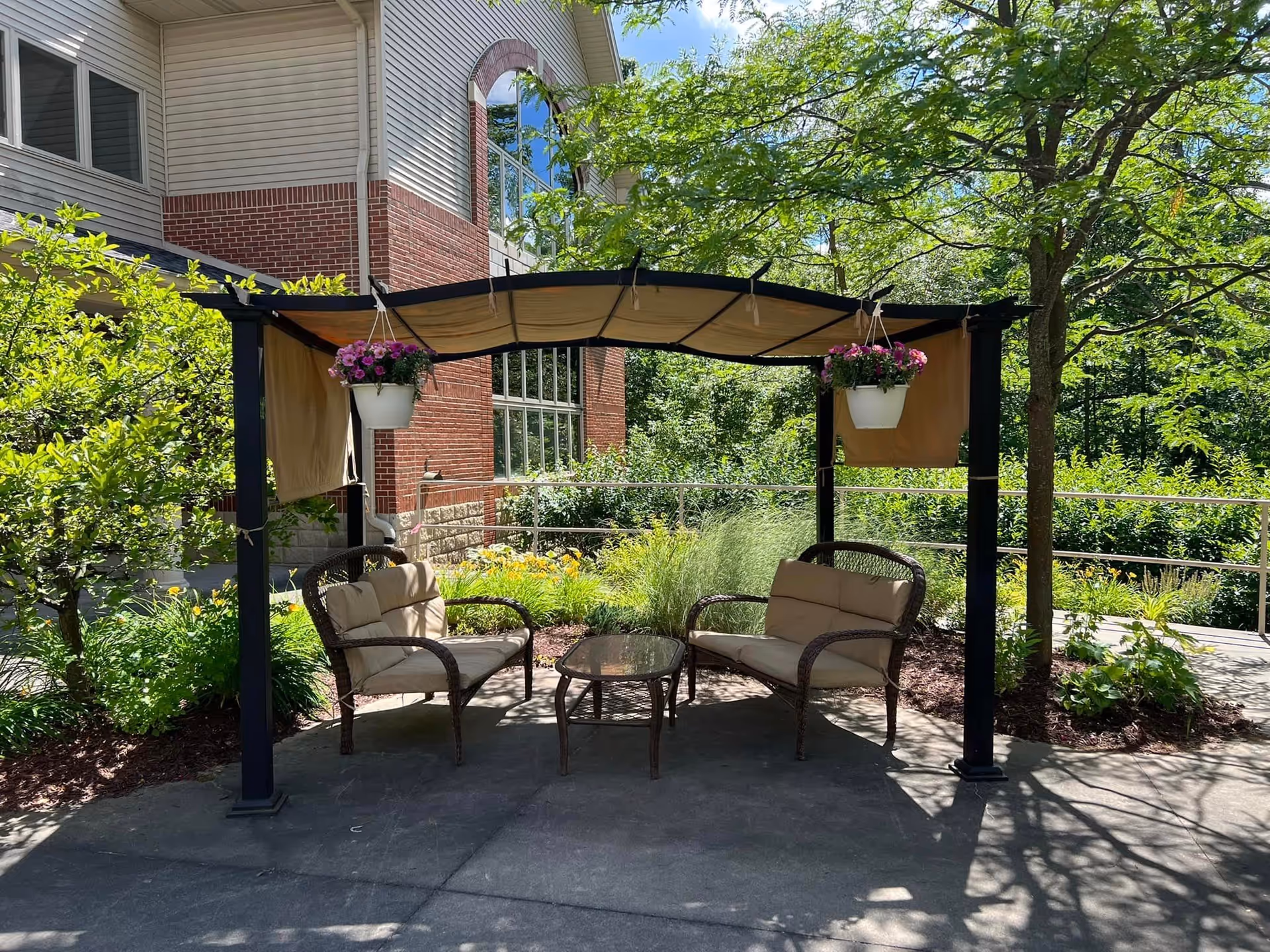 Outdoor seating area with two cushioned chairs and a glass-top table under a pergola with beige fabric canopy and hanging flower pots, surrounded by greenery and trees next to a brick and siding building.