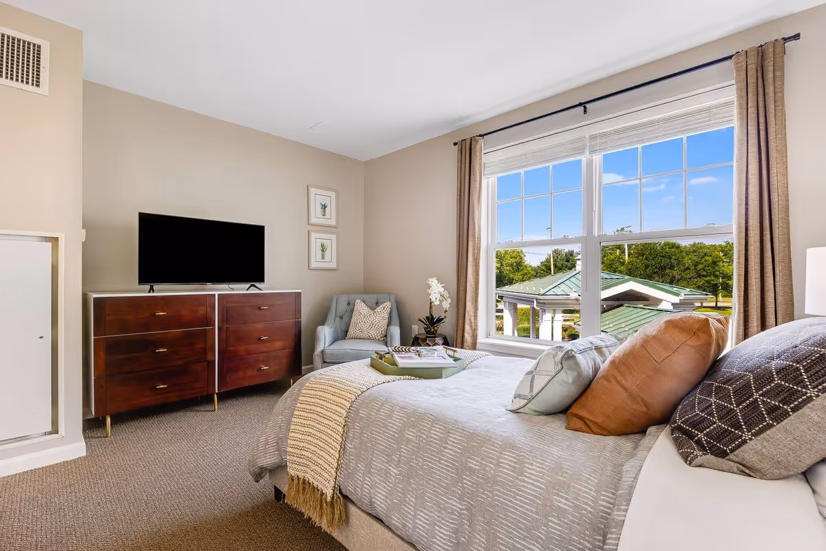 A bright bedroom with a large window showing a view of trees and a green-roofed structure outside. The room features a bed with multiple pillows and a beige throw blanket, a wooden dresser with a flat-screen TV on top, a light blue armchair with a patterned pillow, and two framed botanical prints on the wall.