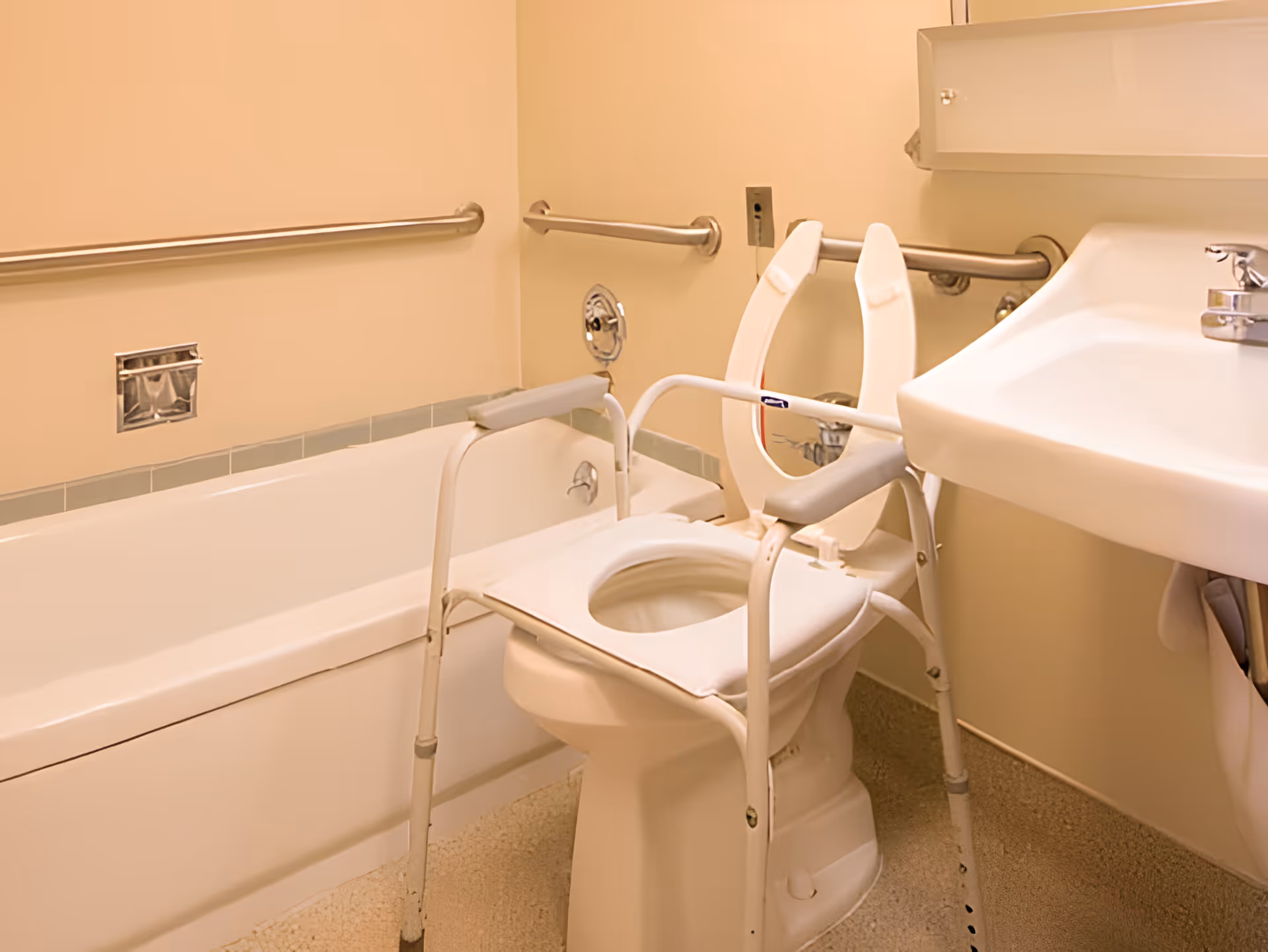 A bathroom with a white bathtub, a white toilet equipped with a raised toilet seat and armrests for accessibility, a white sink with a faucet, and metal grab bars mounted on the walls for support.