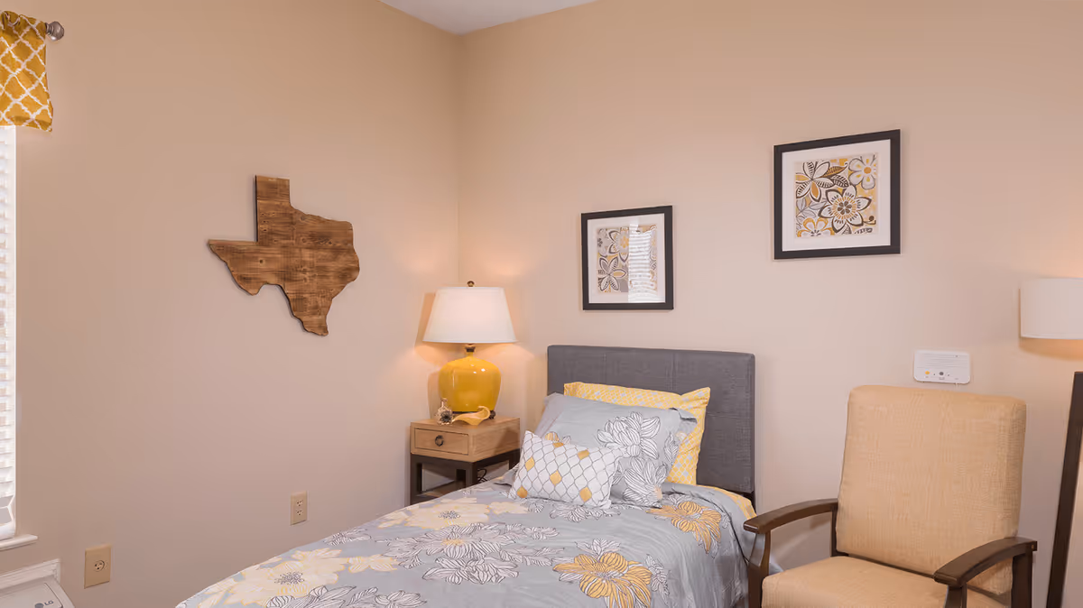 A cozy bedroom with a single bed featuring floral-patterned bedding in shades of gray, yellow, and white. Above the bed are two framed floral artworks. To the left of the bed is a wooden nightstand with a yellow lamp and decorative items. A wooden wall decoration shaped like the state of Texas hangs on the beige wall. To the right of the bed is a beige upholstered armchair with wooden armrests. A window with a yellow patterned valance is partially visible on the left side.