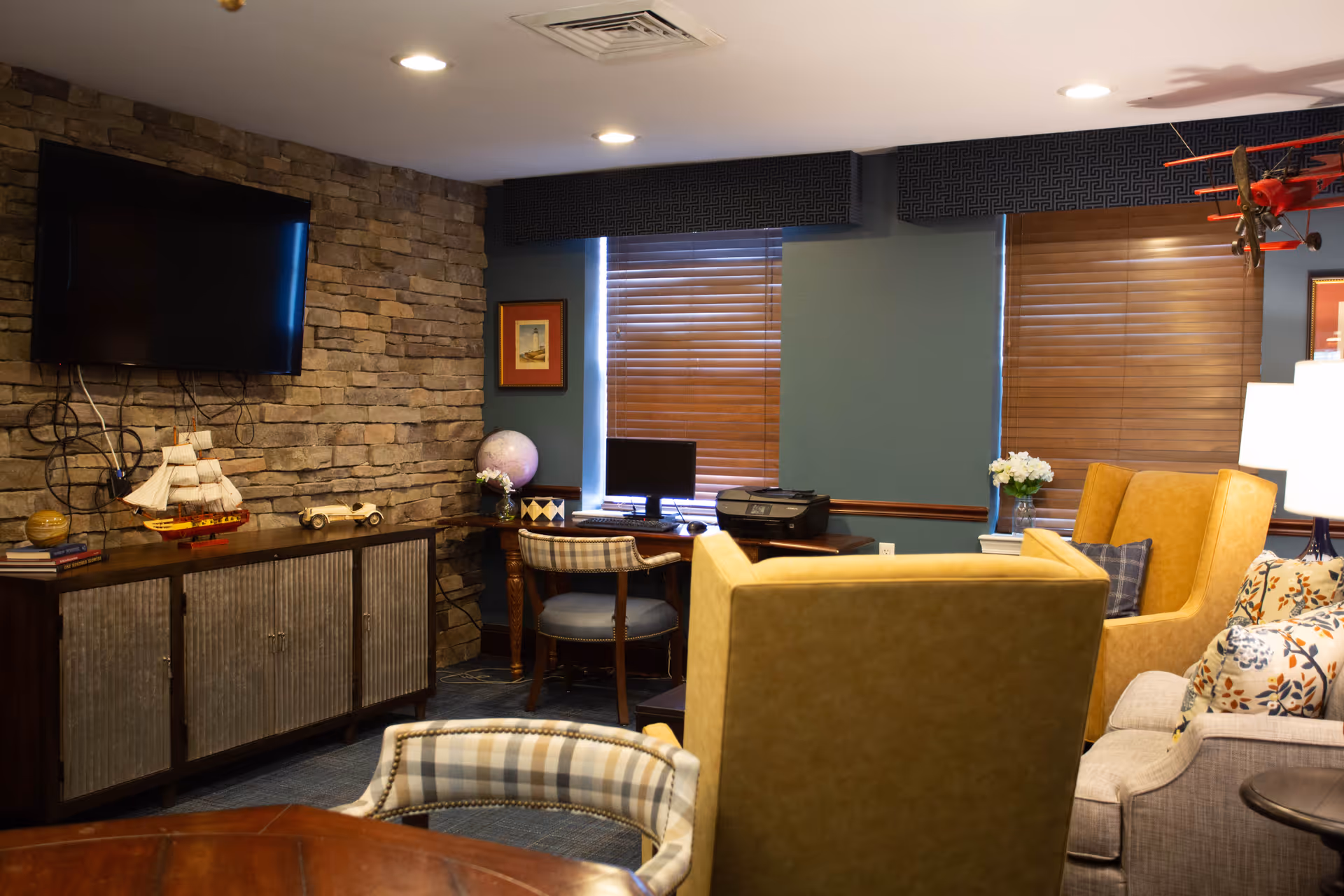 A cozy living room area with a stone accent wall featuring a mounted flat-screen TV. Below the TV is a wooden cabinet with decorative items including a model ship and a small wooden car. The room has blue walls with two windows covered by wooden blinds. There is a desk with a computer and printer, a plaid upholstered chair, and comfortable seating including a yellow armchair and a gray sofa with patterned cushions. A model red airplane hangs from the ceiling near the window.