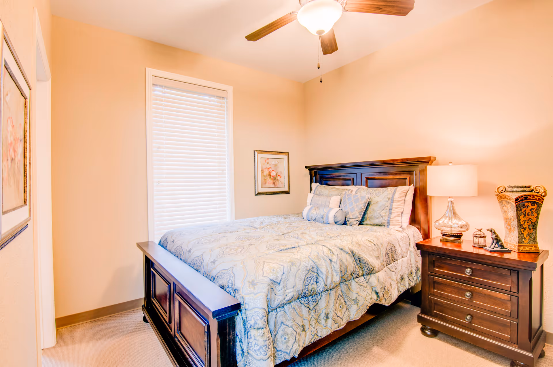 A cozy bedroom with a wooden bed frame and a neatly made bed with patterned bedding and multiple pillows. There is a wooden nightstand with a lamp, decorative vase, and small figurines. The room has beige walls, a window with blinds, a ceiling fan with a light, and framed artwork on the walls.