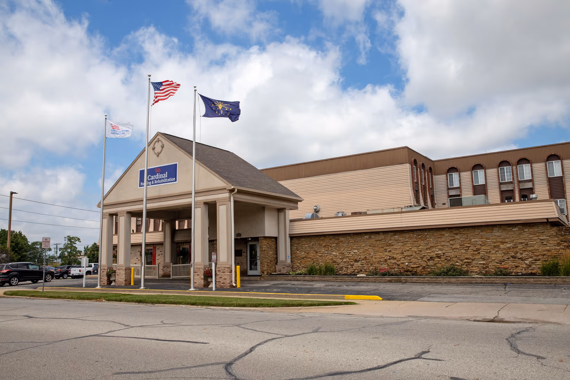 Front entrance of the Cardinal Nursing & Rehabilitation building with three flagpoles flying flags and a parking area under a partly cloudy sky.
