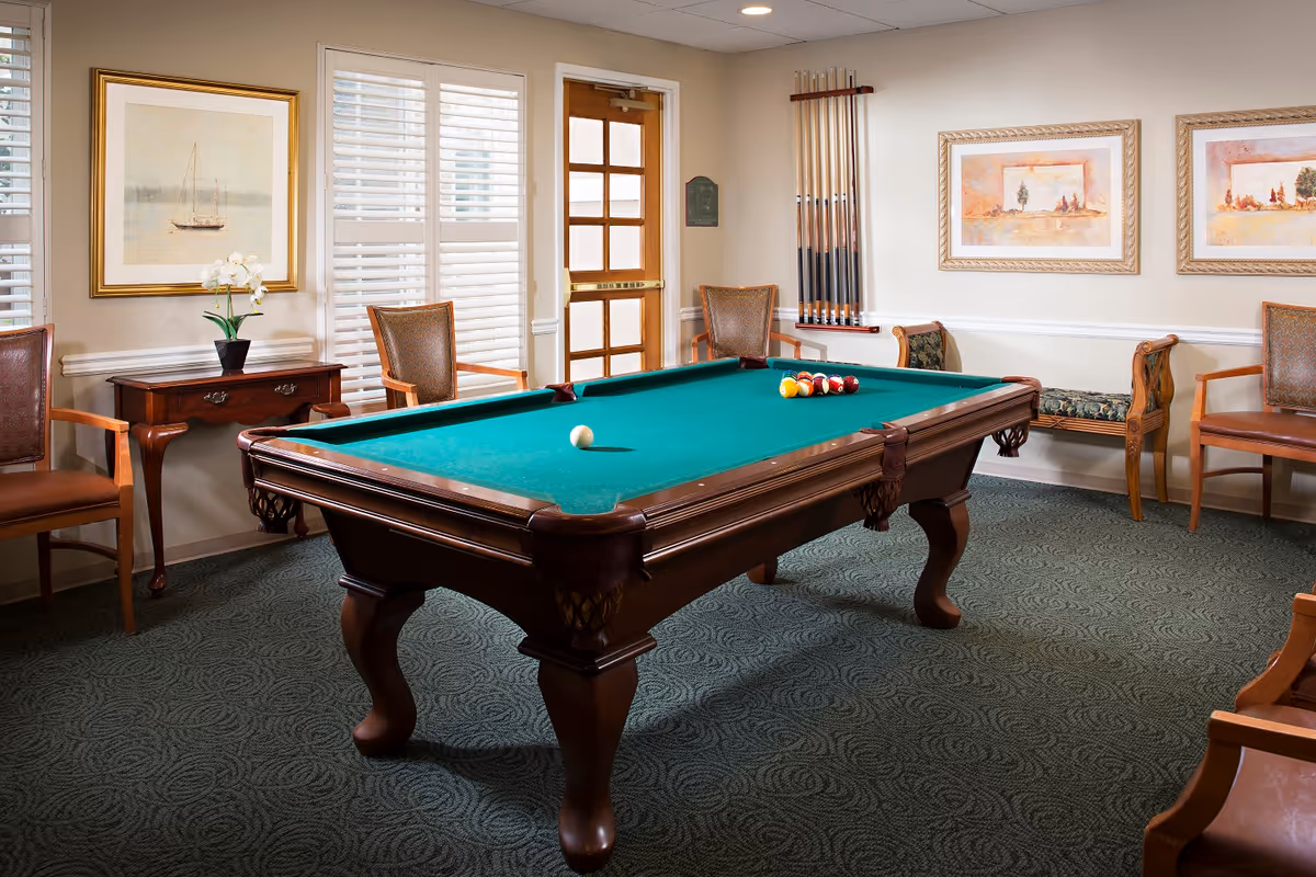 Billiards table in a furnished game room with chairs, framed artwork, and windows with shutters.