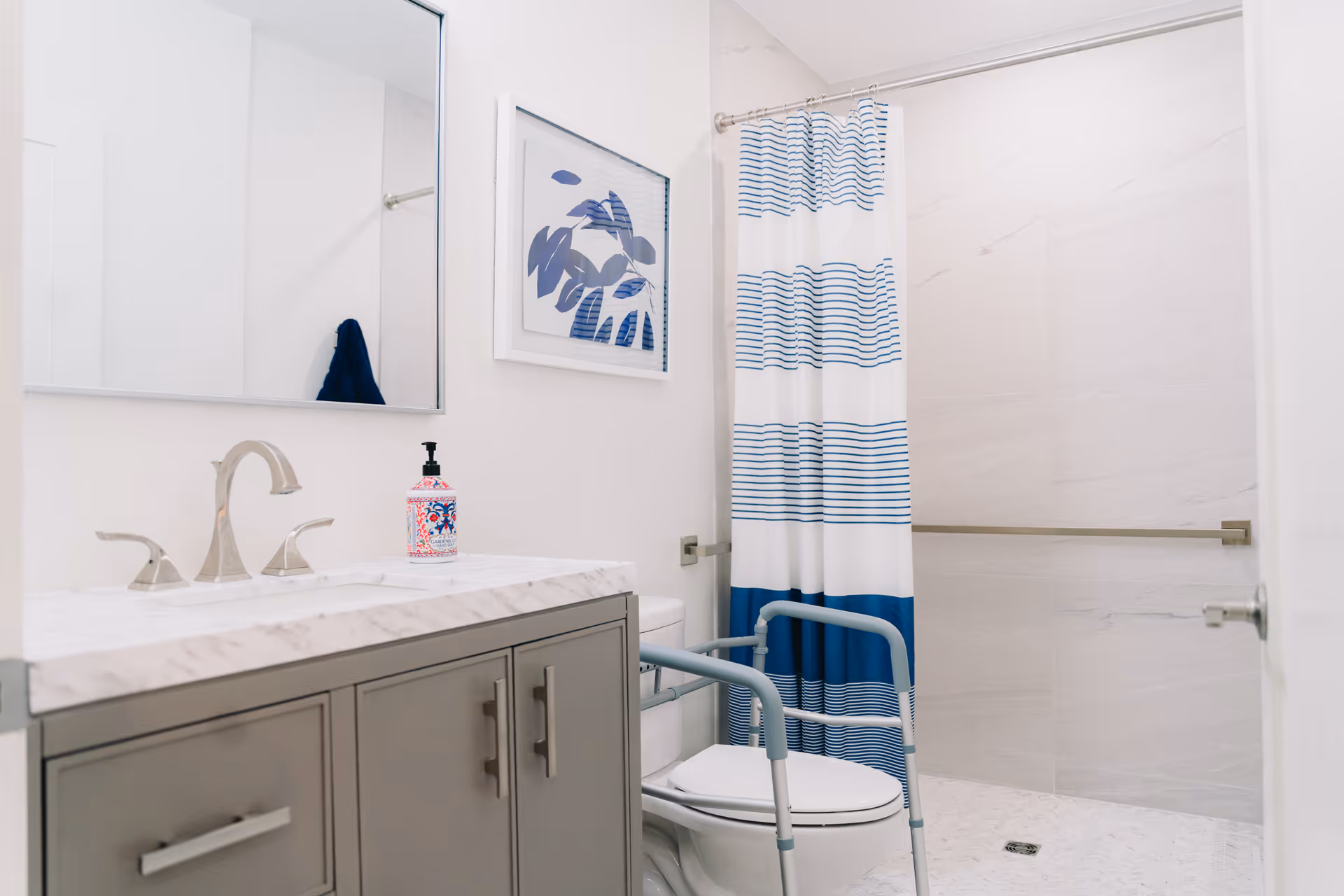 Bright bathroom with a marble-top vanity and mirror, a toilet fitted with a blue mobility frame and grab bar, and a shower behind a blue-striped curtain.