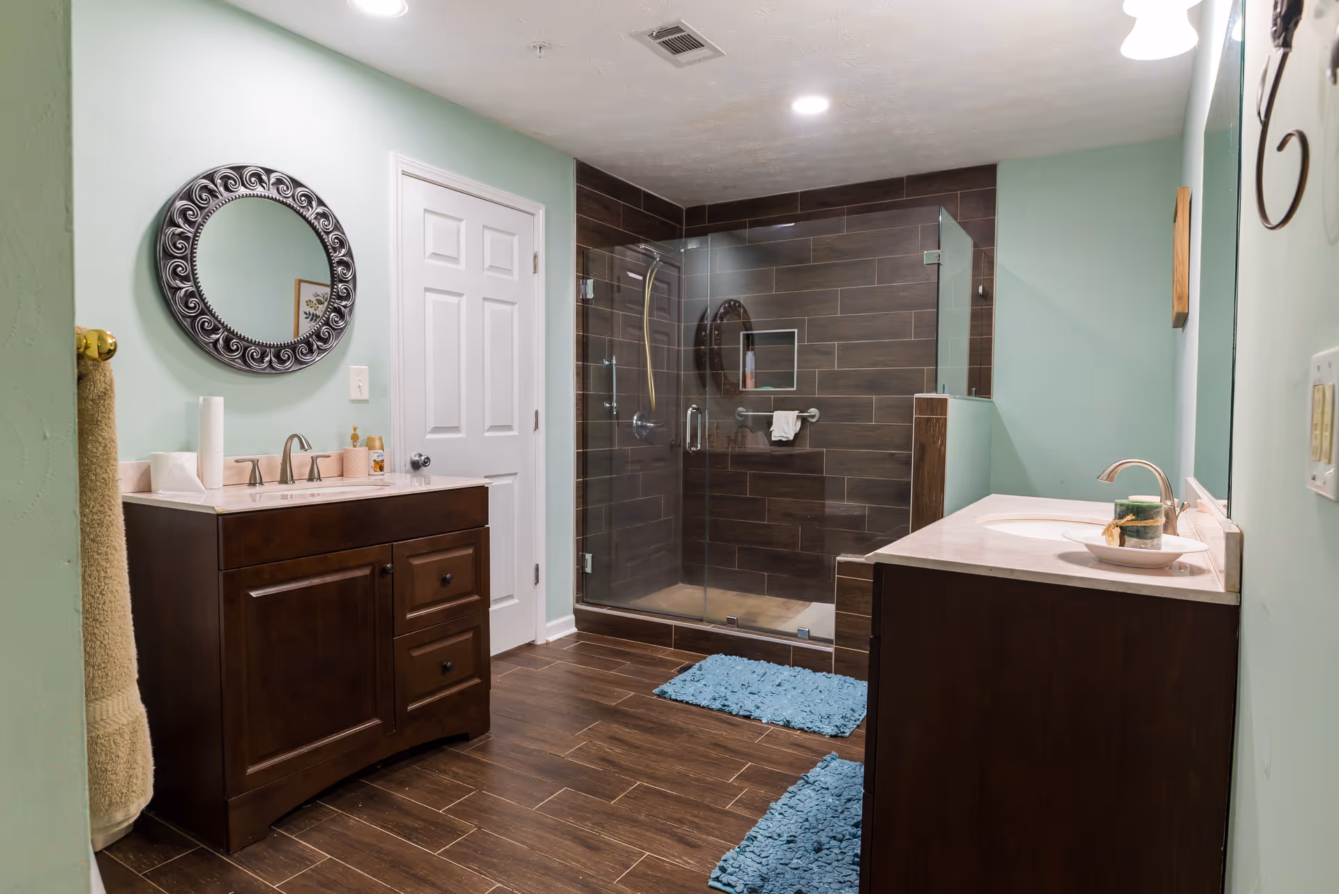 A modern bathroom with light green walls, two dark wood vanities with sinks and mirrors above them, a glass-enclosed shower with dark brown tiles, and two blue bath mats on a wood-look tile floor.