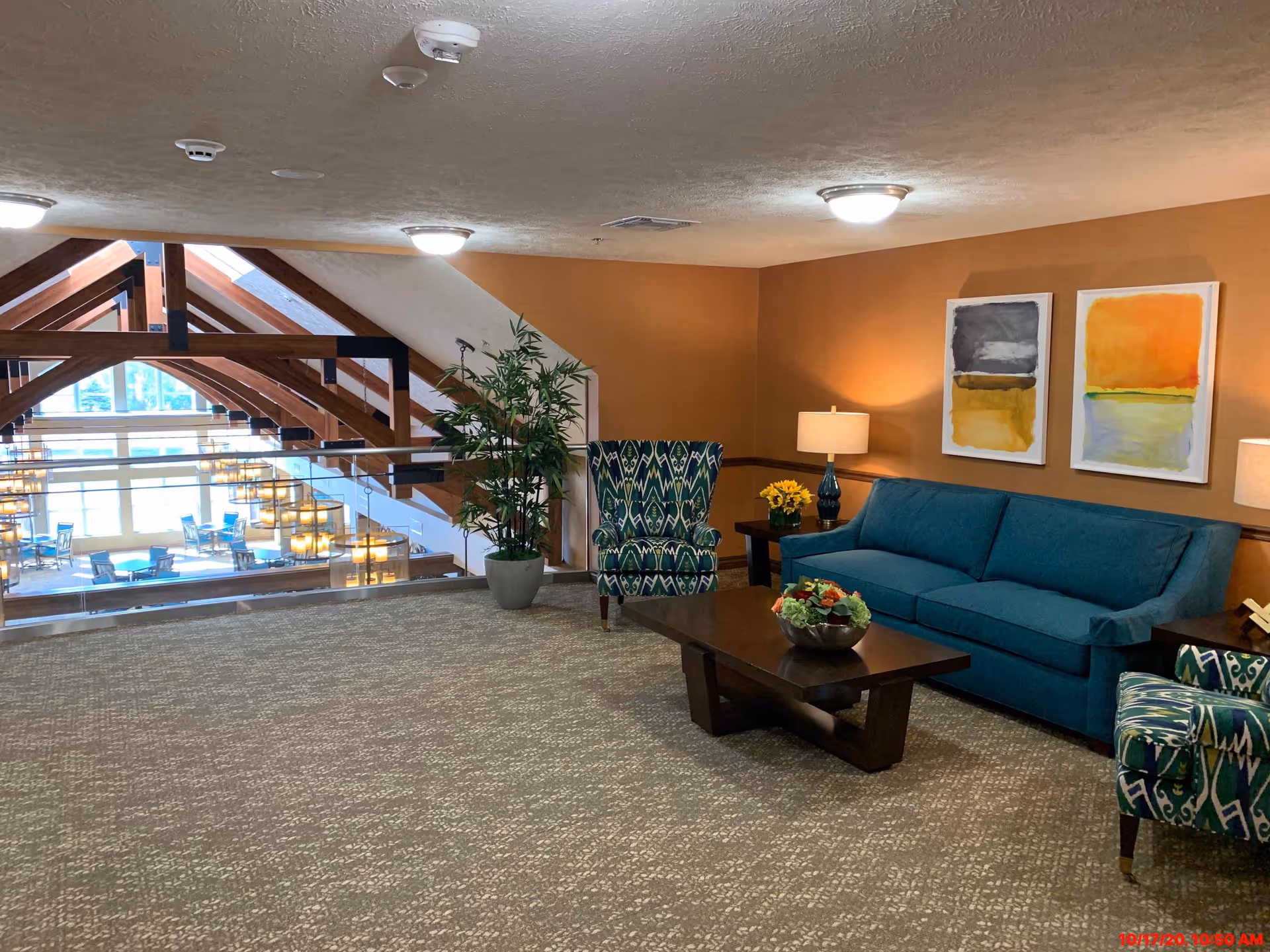 A cozy seating area in a retirement resort with a teal sofa, two patterned armchairs, a dark wooden coffee table with a floral arrangement, two side tables with lamps, and two abstract paintings on an orange wall. The area overlooks a large open space with wooden beams and glass railings.