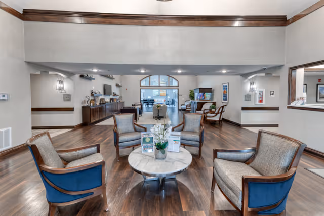 A spacious senior living facility common area with four cushioned armchairs arranged around a round marble coffee table with a small plant and informational brochures. The room features wood flooring, neutral-colored walls with dark wood trim, and recessed lighting. In the background, there is a long cabinet with decorative items and a large window letting in natural light.