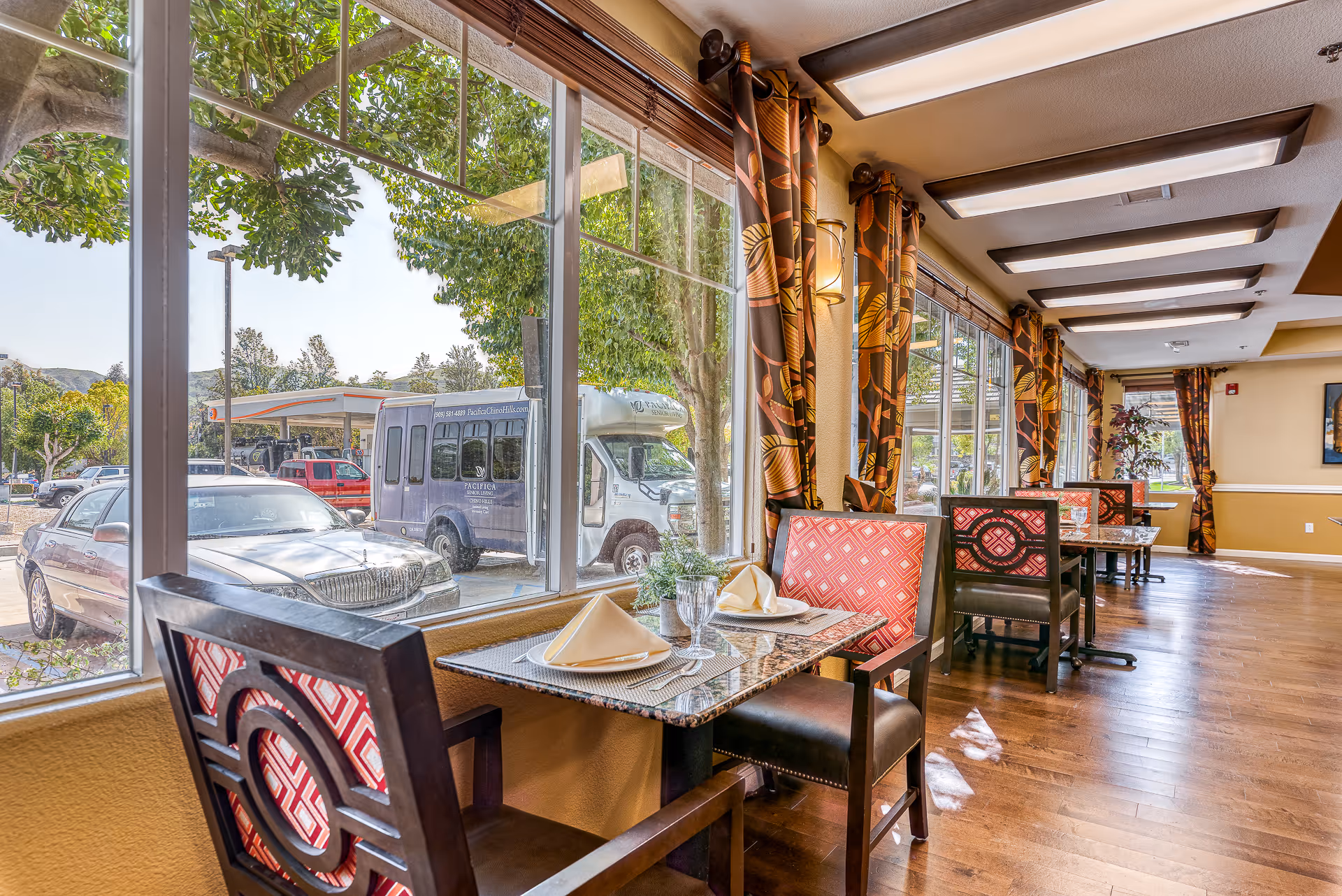 Dining area with tables and chairs set near large windows showing a parking lot outside. Tables have place settings with napkins and glassware. The room has wooden floors, patterned curtains, and ceiling lights.