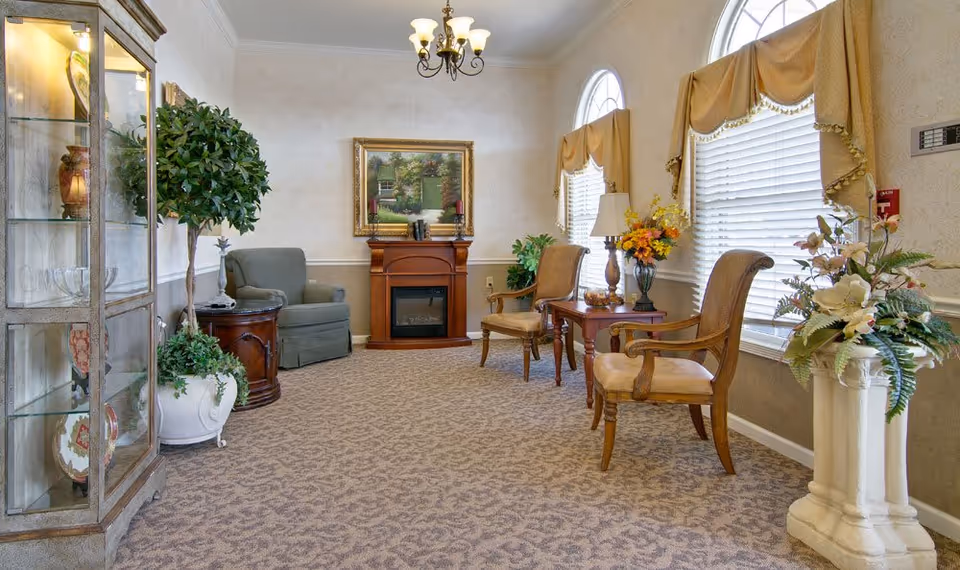 A cozy senior living facility common area with beige patterned carpet, two wooden armchairs with cushions, a small wooden table with a lamp and flower arrangement, a green armchair next to a round wooden side table, a fireplace with a framed painting above it, large windows with beige valances, and decorative plants including a tall potted tree and a floral arrangement on a pedestal.