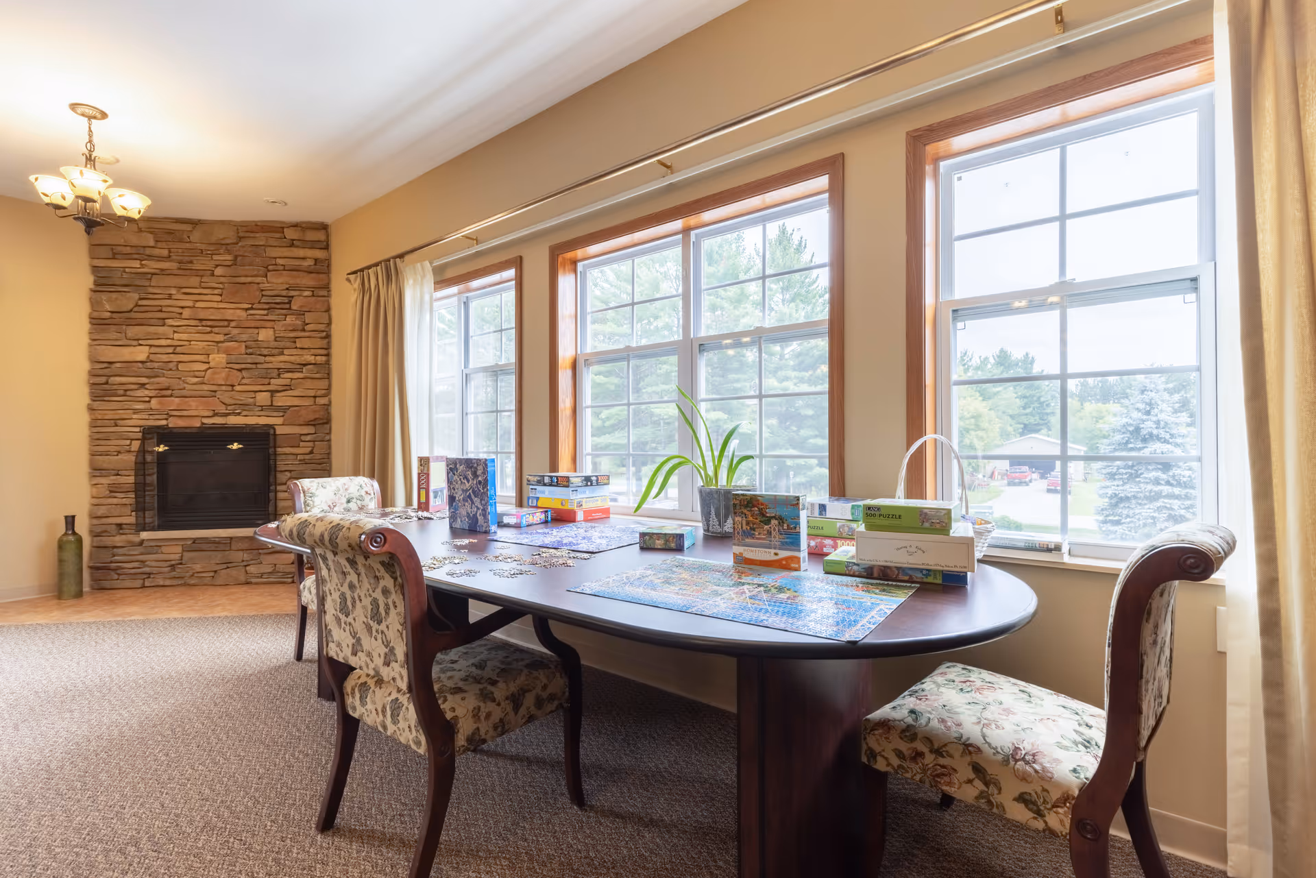 Sunlit common room with a large table covered in puzzle pieces and boxes, floral-upholstered chairs, a stone fireplace, and tall windows overlooking greenery.