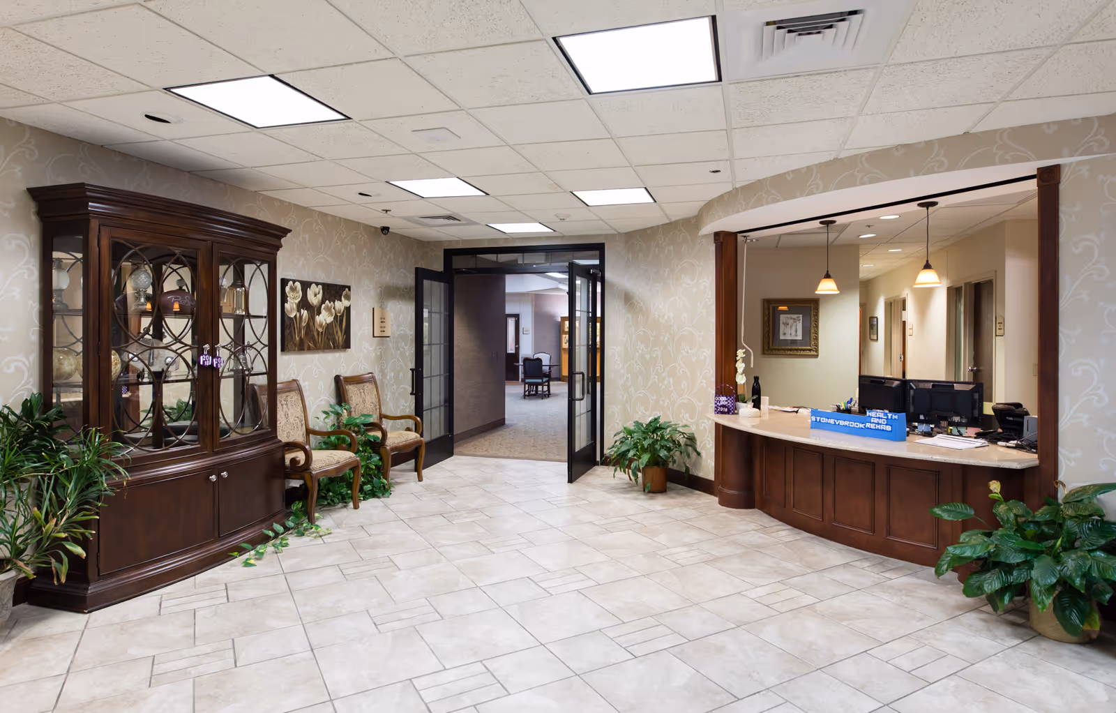 Bright lobby reception area with a wooden front desk, seating, display cabinet, plants, and open double doors to a hallway.