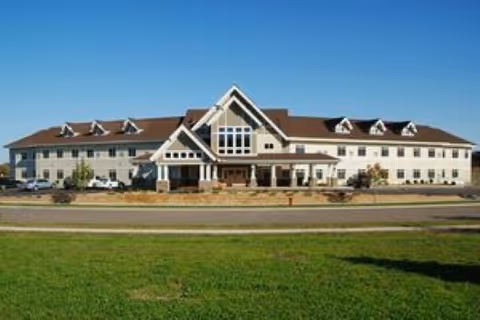 Large two-story beige senior living building with a central peaked entrance, columns, and a grassy lawn under a clear blue sky.