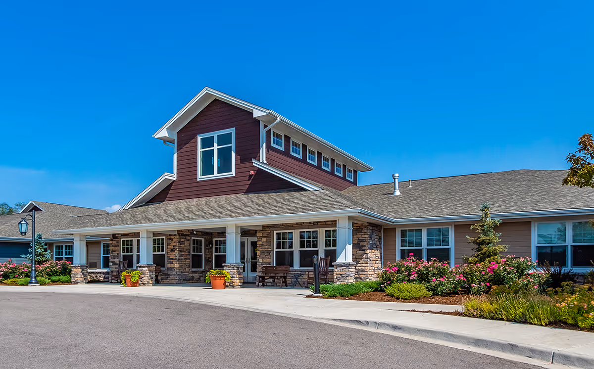 Exterior view of a single-story senior living facility building with stone and wood siding, large windows, a covered entrance with white columns, potted plants, benches, and landscaped flower beds under a clear blue sky.