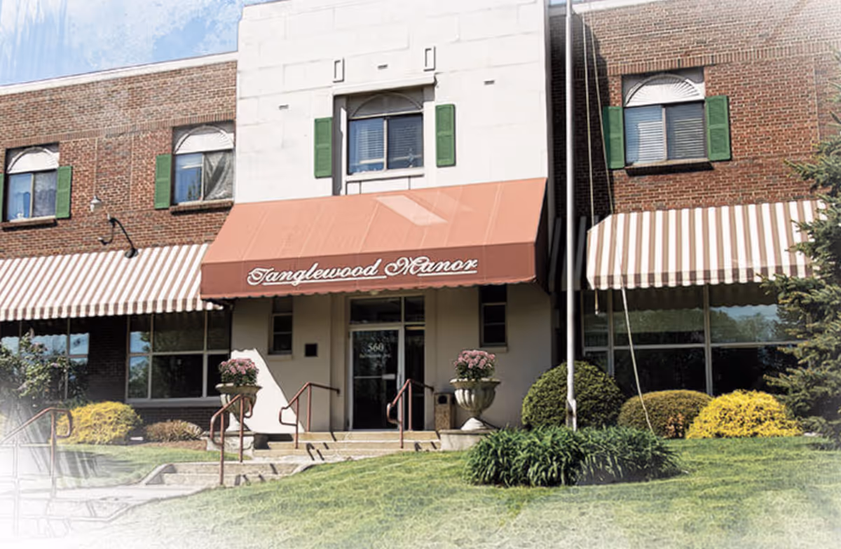 Front entrance of Tanglewood Manor with a red awning showing the facility name, flanked by windows and landscaped shrubs.