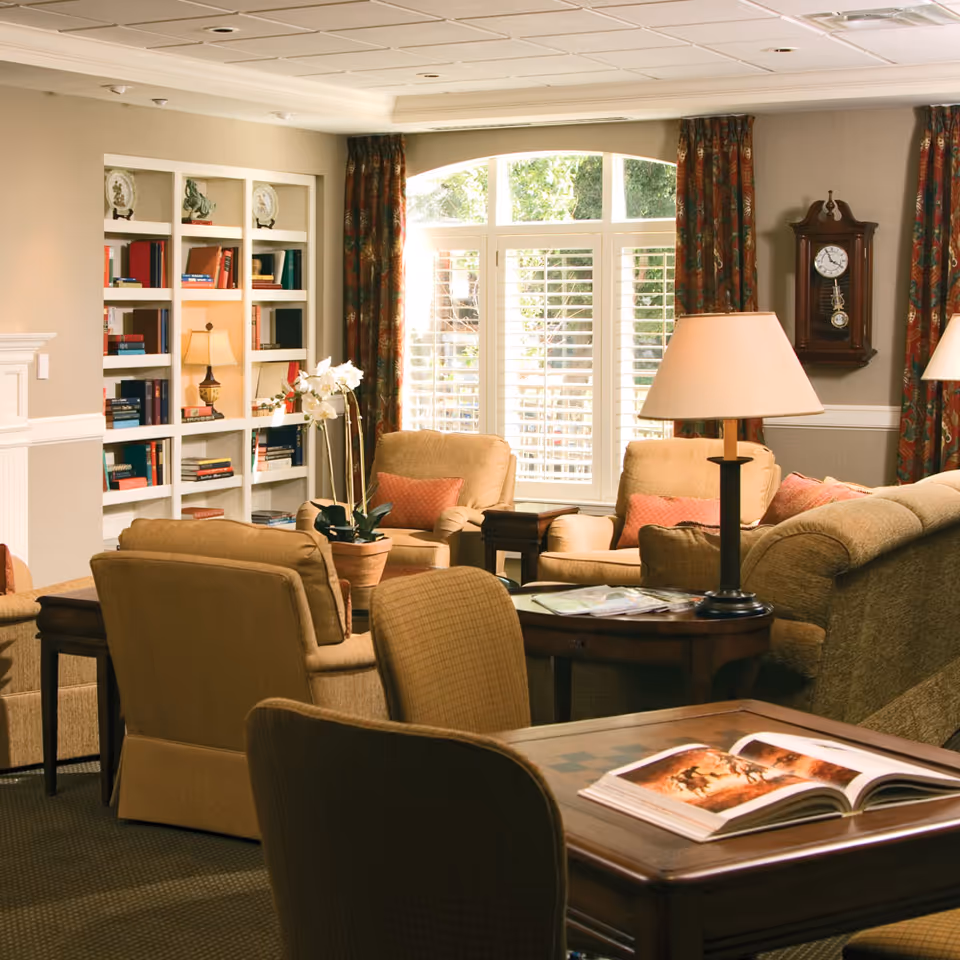 Cozy living room with upholstered chairs and sofas, a bookshelf, table lamps, and a large window with shutters.
