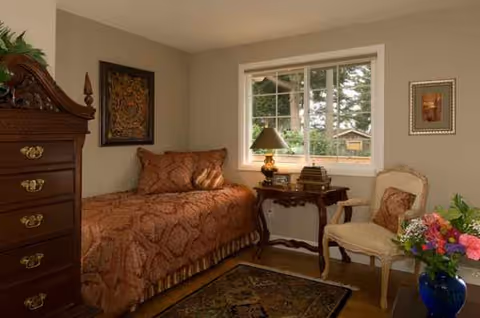 Cozy bedroom with a single bed, dresser, nightstand lamp, window, chair, and flower vase.