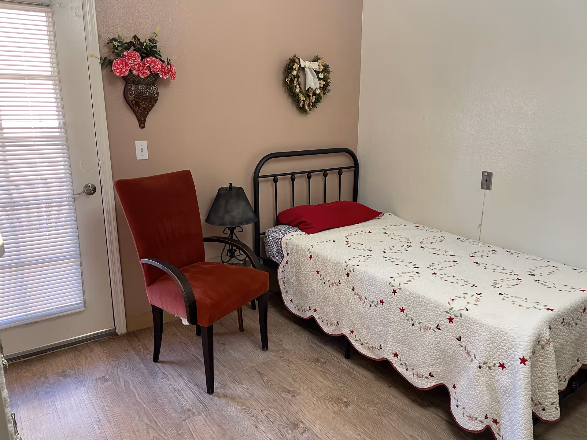 Small bedroom with a single metal-framed bed covered in a white quilt, a red chair, bedside lamp, and floral wall decorations by a door with blinds.