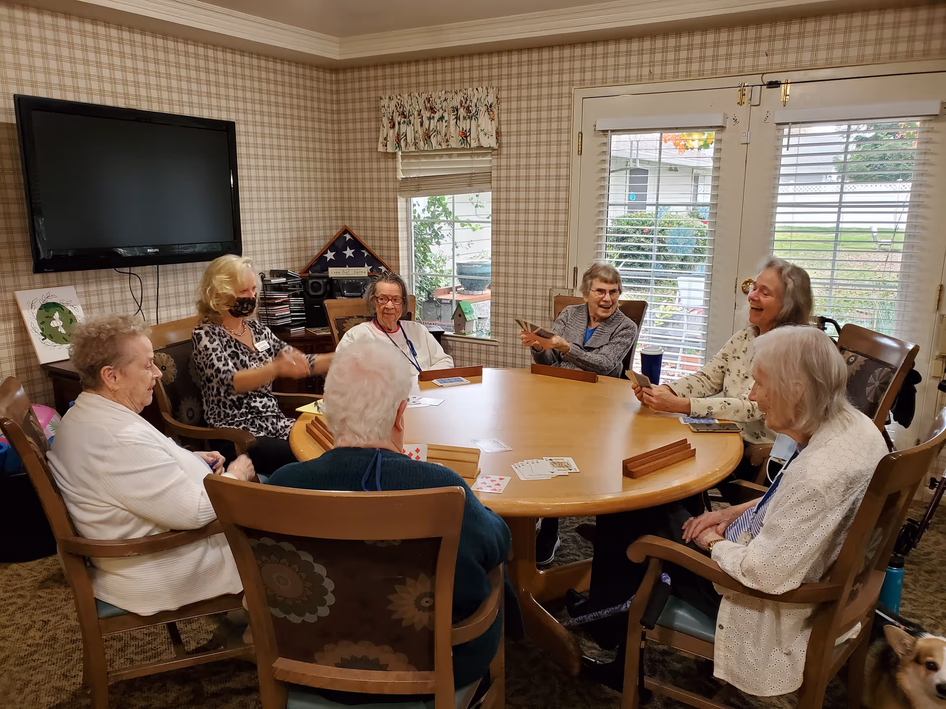 A group of elderly women sitting around a large round wooden table playing cards in a cozy room with plaid wallpaper, a wall-mounted TV, and windows with blinds and floral valances. One woman is wearing a face mask, and a dog is partially visible in the bottom right corner.