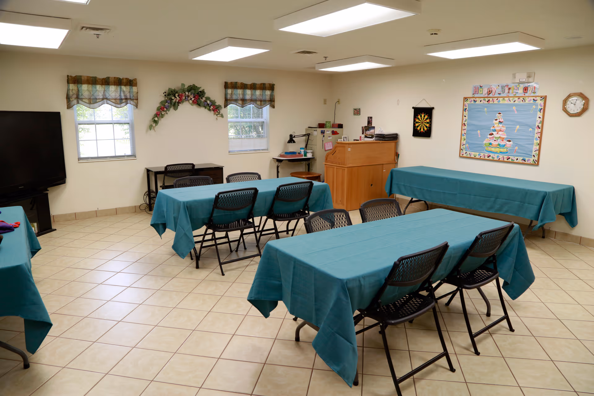 Community activity/dining room with tables covered in teal tablecloths, folding chairs, a TV, and a bulletin board on a tiled floor.