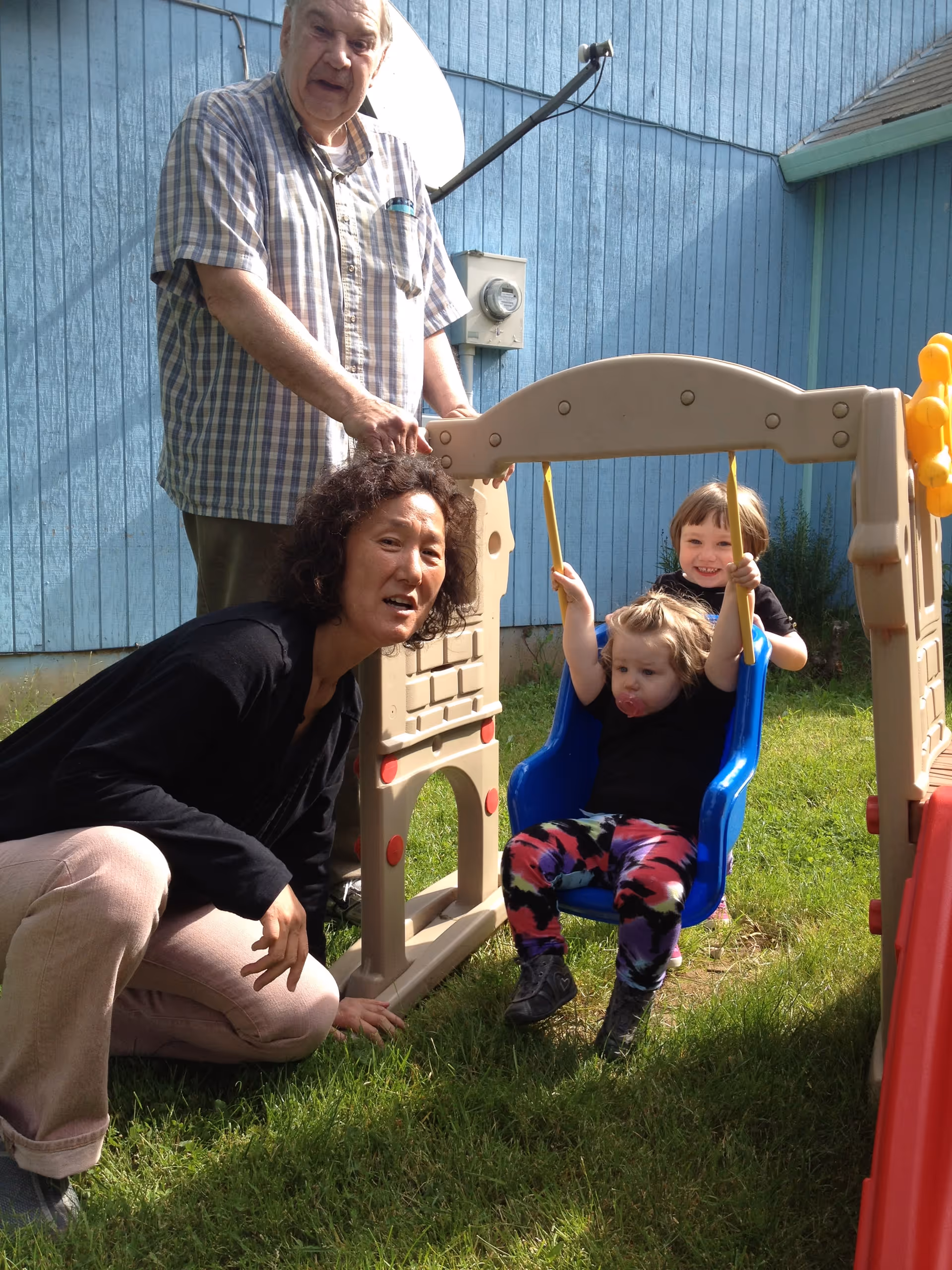 An elderly man and a woman are outside near a blue wooden building. The man is standing behind a small plastic swing set, while the woman is crouching beside it. Two young children are playing on the swing; one child is sitting on the swing and the other is standing behind it, smiling.