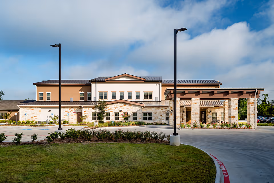 Front exterior view of a two-story senior living facility building with stone and beige siding, a covered entrance supported by stone pillars, landscaped greenery, and a driveway with street lamps under a partly cloudy sky.