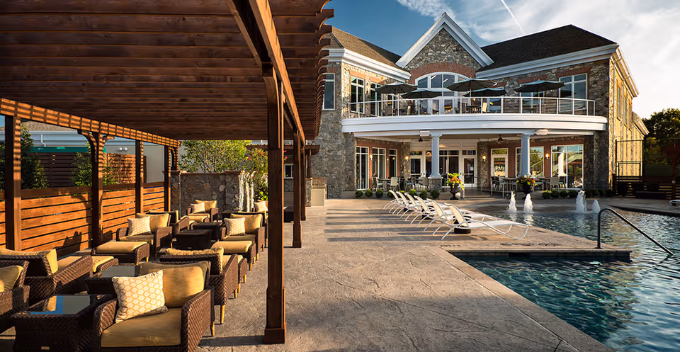 Outdoor seating area with cushioned wicker chairs under a wooden pergola next to a swimming pool with lounge chairs and water fountains, in front of a large stone building with a balcony and patio furniture.