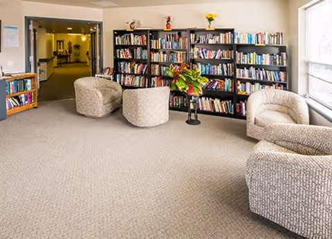 A cozy reading area in a senior living facility with three upholstered armchairs arranged around a small round table holding a flower arrangement. Behind the chairs are bookshelves filled with books. The room has beige carpeting and a large window on the right side letting in natural light. An open doorway leads to another part of the facility.