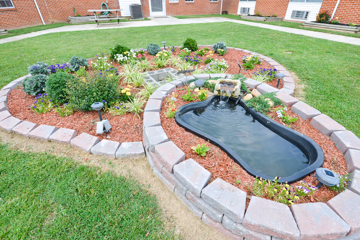 A landscaped garden area with a small pond surrounded by stone bricks and mulch, featuring various plants and flowers. The garden is set within a grassy courtyard area bordered by brick buildings with windows and a door visible in the background.