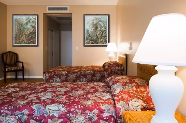 A bedroom with two single beds covered in red floral bedspreads. There is a wooden nightstand with a white lamp between the beds. On the wall behind the beds are two framed botanical prints. A wooden chair with striped upholstery is placed against the far wall near an open doorway.
