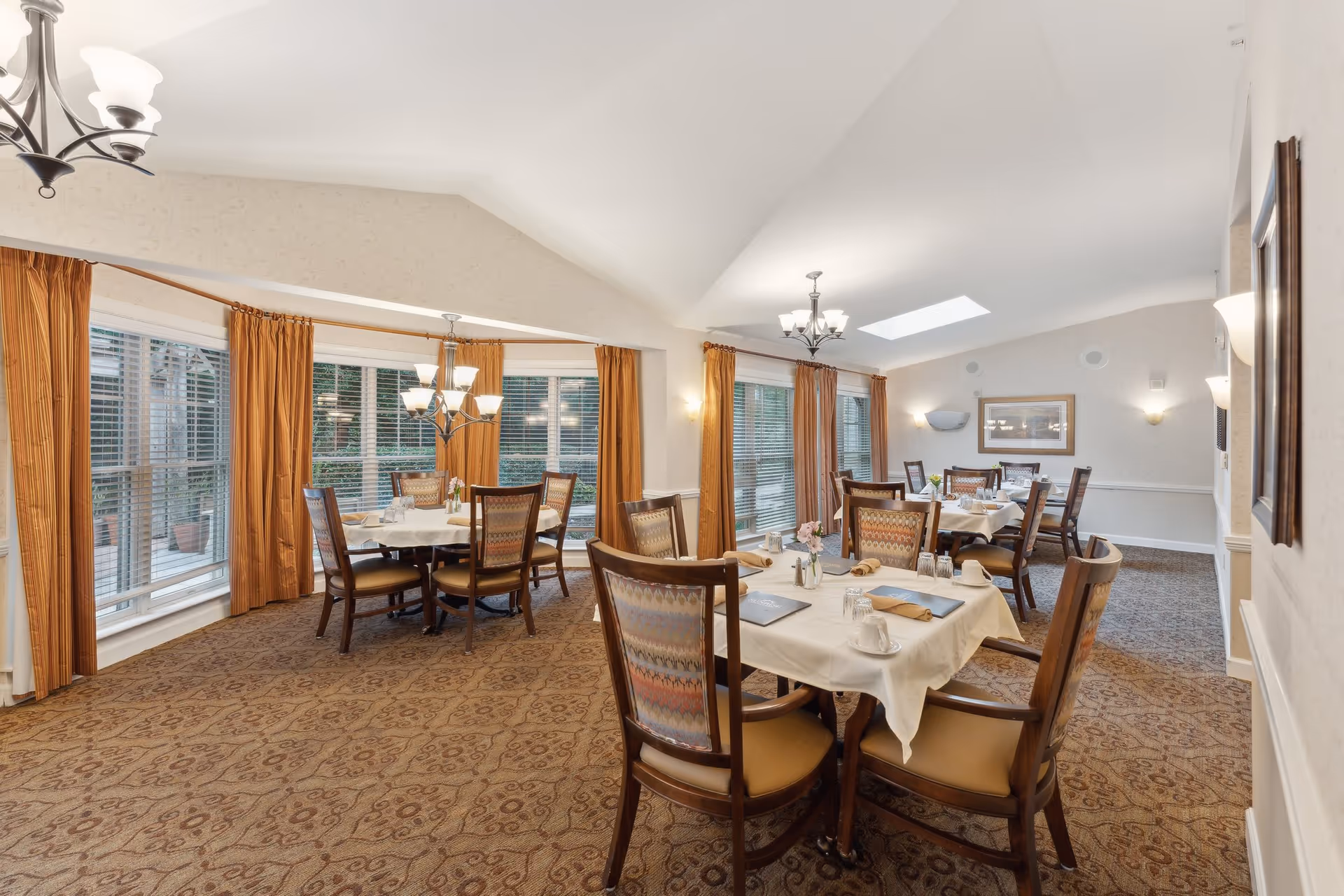 A bright dining room in an assisted living facility with multiple round tables covered with white tablecloths, set with plates, cups, and napkins. The room has large windows with orange curtains, patterned carpet, and chandeliers hanging from a vaulted ceiling.