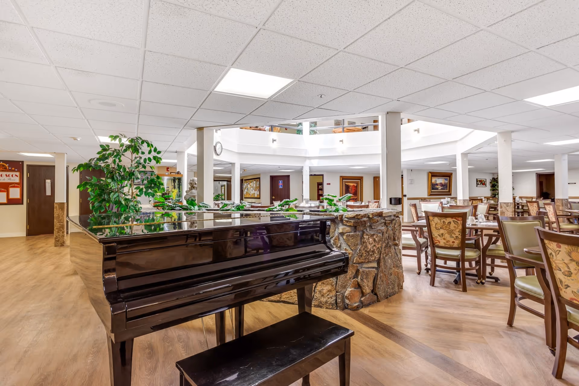 Interior view of a senior living community dining area featuring a black grand piano in the foreground, wooden tables and chairs with floral upholstery, a stone planter with green plants, and a bright ceiling with recessed lighting.