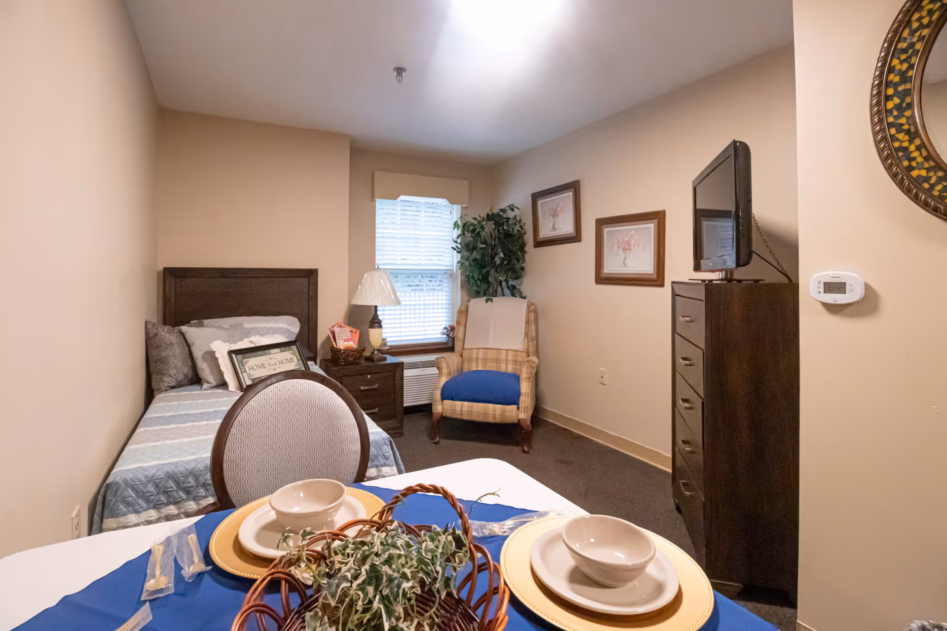 A small bedroom with a single bed against the left wall, a nightstand with a lamp next to the bed, a cushioned armchair near a window with blinds, a tall dresser with a TV on top, and a dining table set with plates, bowls, and a centerpiece in the foreground.