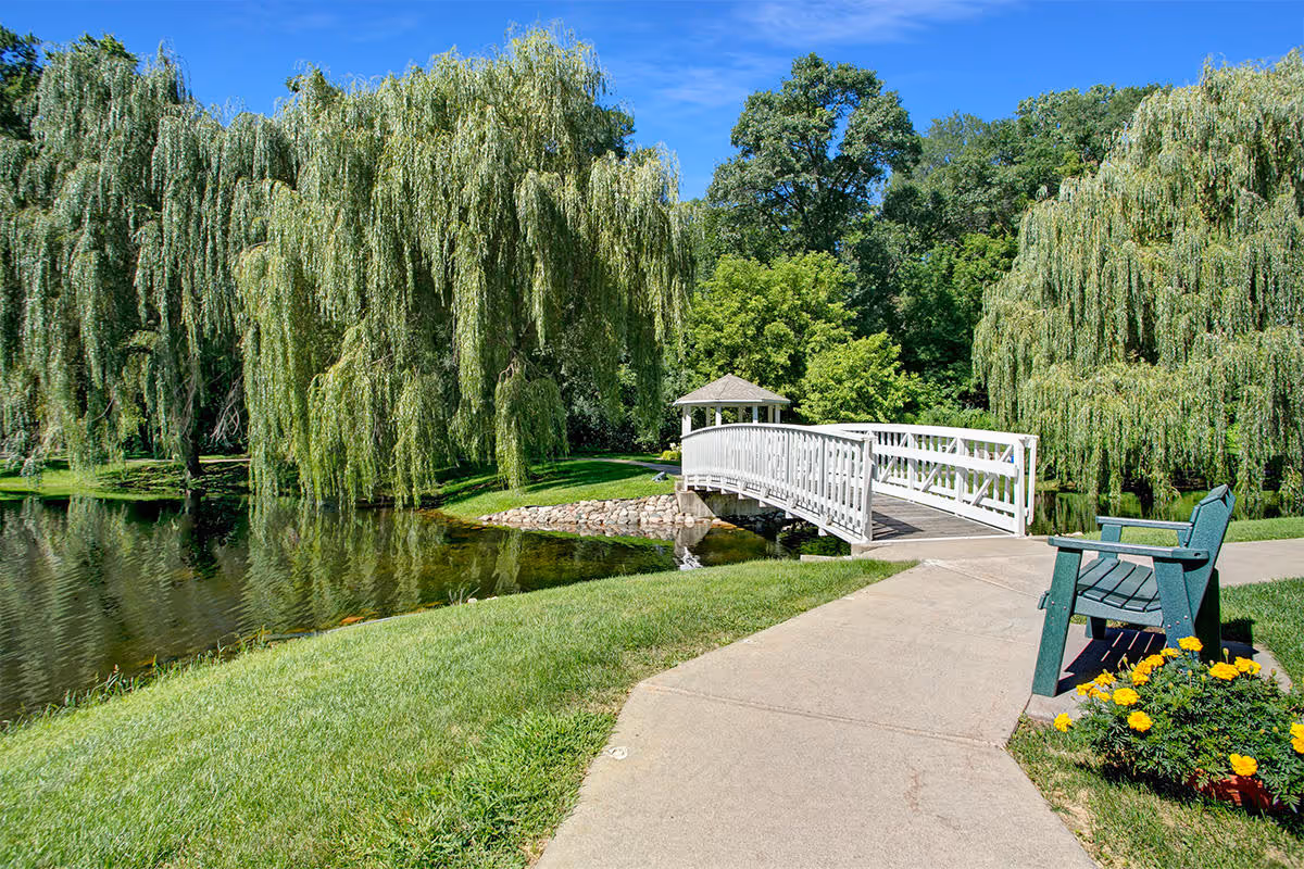 A white footbridge over a pond surrounded by willow trees, with a paved path, bench, and flowers in the foreground.