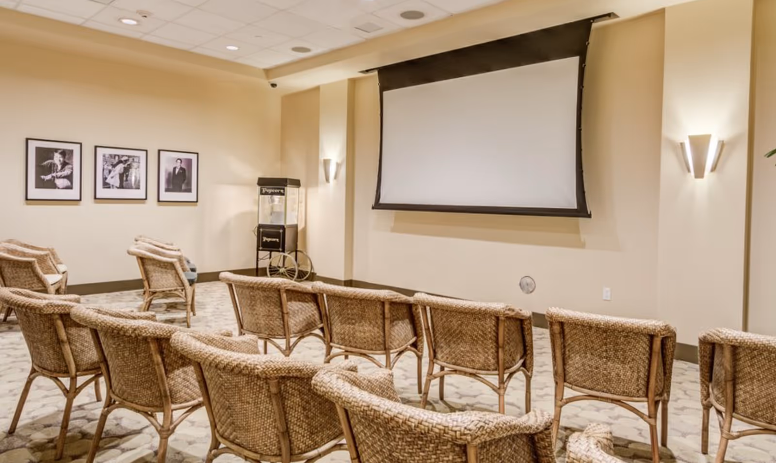 A small theater room with rows of wicker chairs facing a large blank projector screen. The room has beige walls with three framed black and white photographs on one wall and a popcorn machine in the corner. The floor is carpeted with a patterned design and there are wall-mounted lights providing illumination.