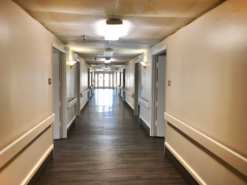 A long, well-lit hallway in an assisted living facility with beige walls, wooden flooring, handrails on both sides, and multiple open doorways leading to rooms. The hallway ends with glass doors letting in natural light.