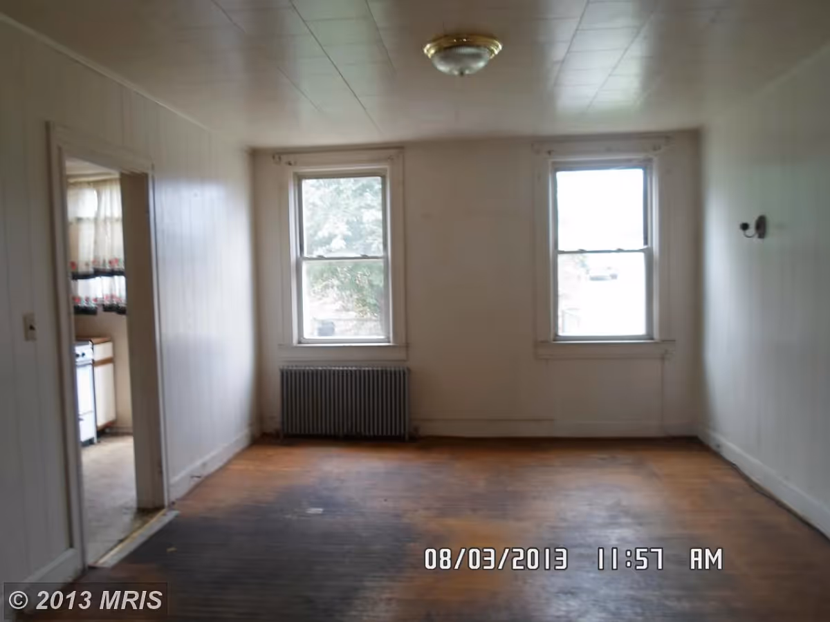 Empty room with two windows, a radiator below one window, wooden floor, white paneled walls, and a ceiling light fixture. There is an open doorway on the left side leading to a kitchen area with a stove and curtains on the window.