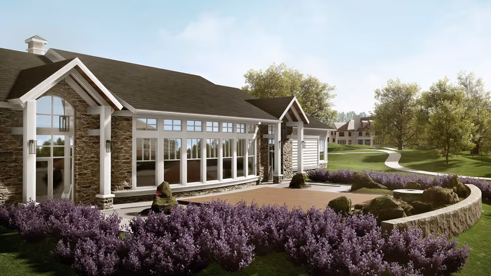 Exterior view of a senior living facility building with large windows, stone and white siding walls, surrounded by a landscaped garden with purple flowering bushes and green trees under a clear sky.