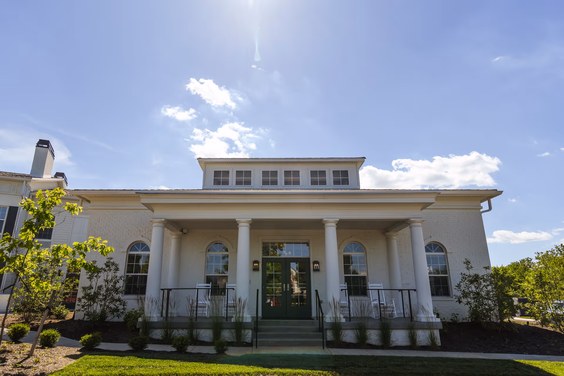 Front exterior view of a white building with large columns, green double doors, and several windows under a clear blue sky with some clouds. There are small bushes and trees planted around the building and a well-maintained lawn in front.