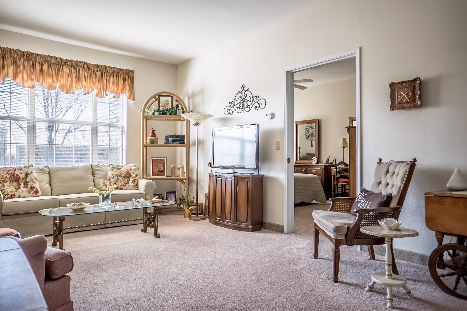 A bright and cozy living room with a large window covered by a valance, a beige sofa with floral cushions, a glass coffee table, a wooden TV stand with a flat-screen TV, a tall shelf with decorative items, a cushioned armchair with a pillow, a small round side table, and a view into a bedroom with a wooden dresser and mirror.