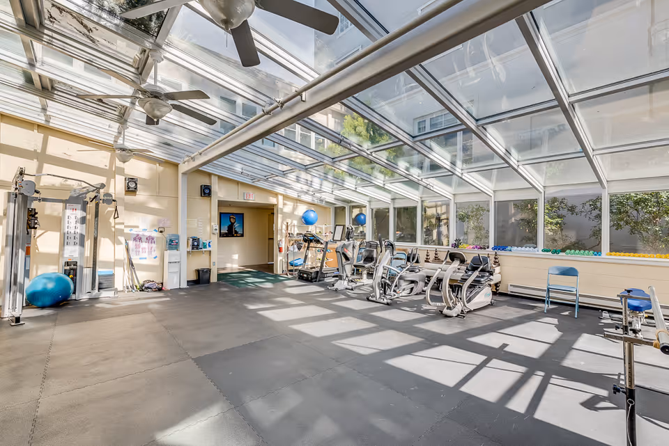 Sunlit indoor fitness room with exercise machines, weights, and a glass skylight ceiling.