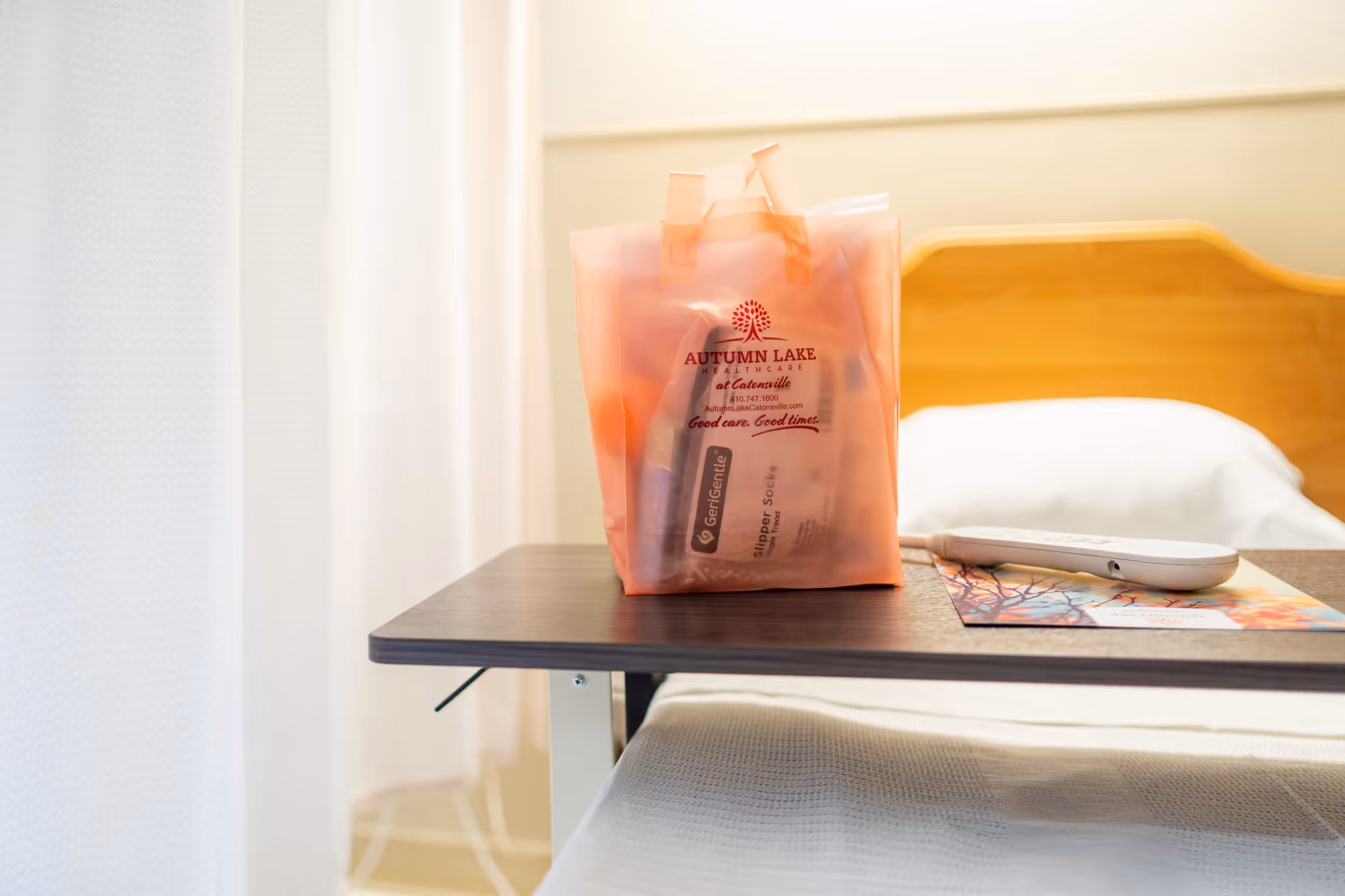 A bedside table with a translucent orange bag containing medical supplies, a remote control, and a colorful brochure, next to a hospital-style bed with white bedding and a wooden headboard.